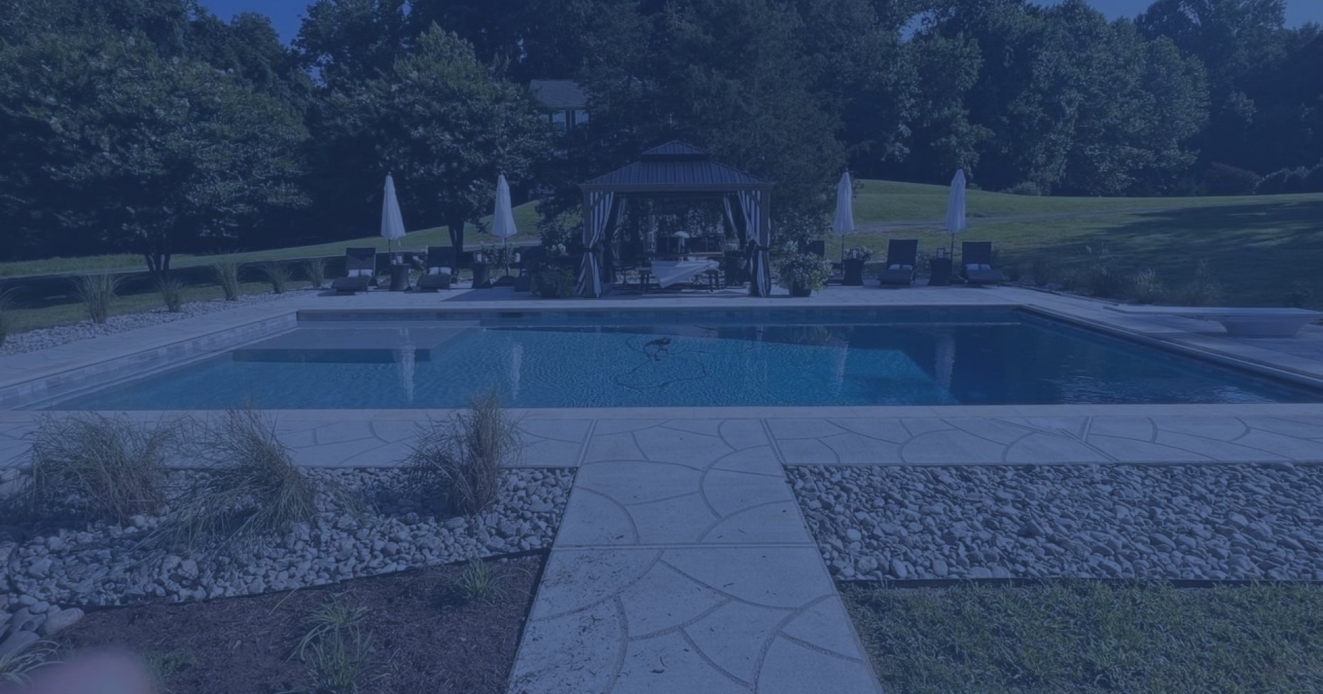 A landscaped backyard patio beside a modern house at dusk with a pool, fire pit seating, and warm interior lighting.